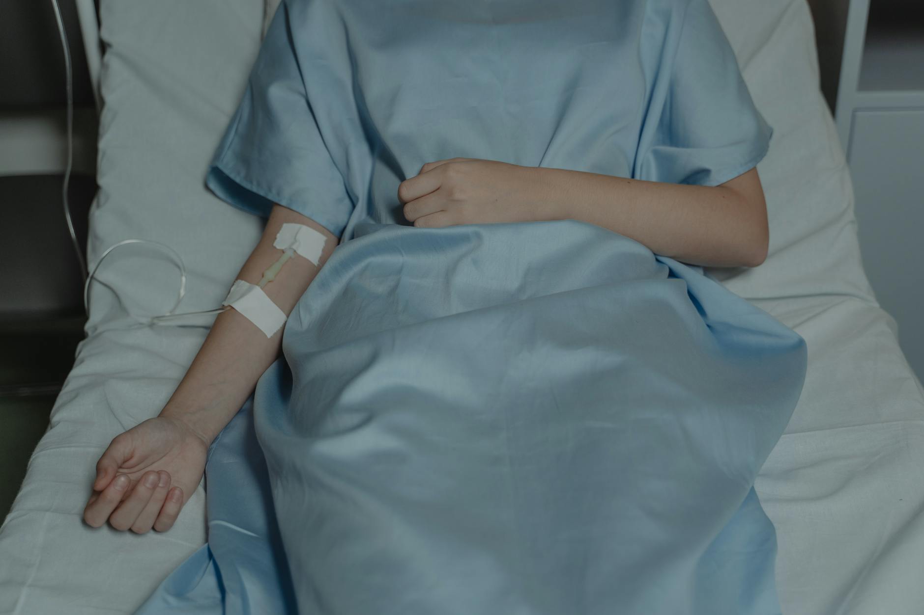 A patient lies in bed wearing a hospital gown with an IV drip attached, evoking a sense of illness.