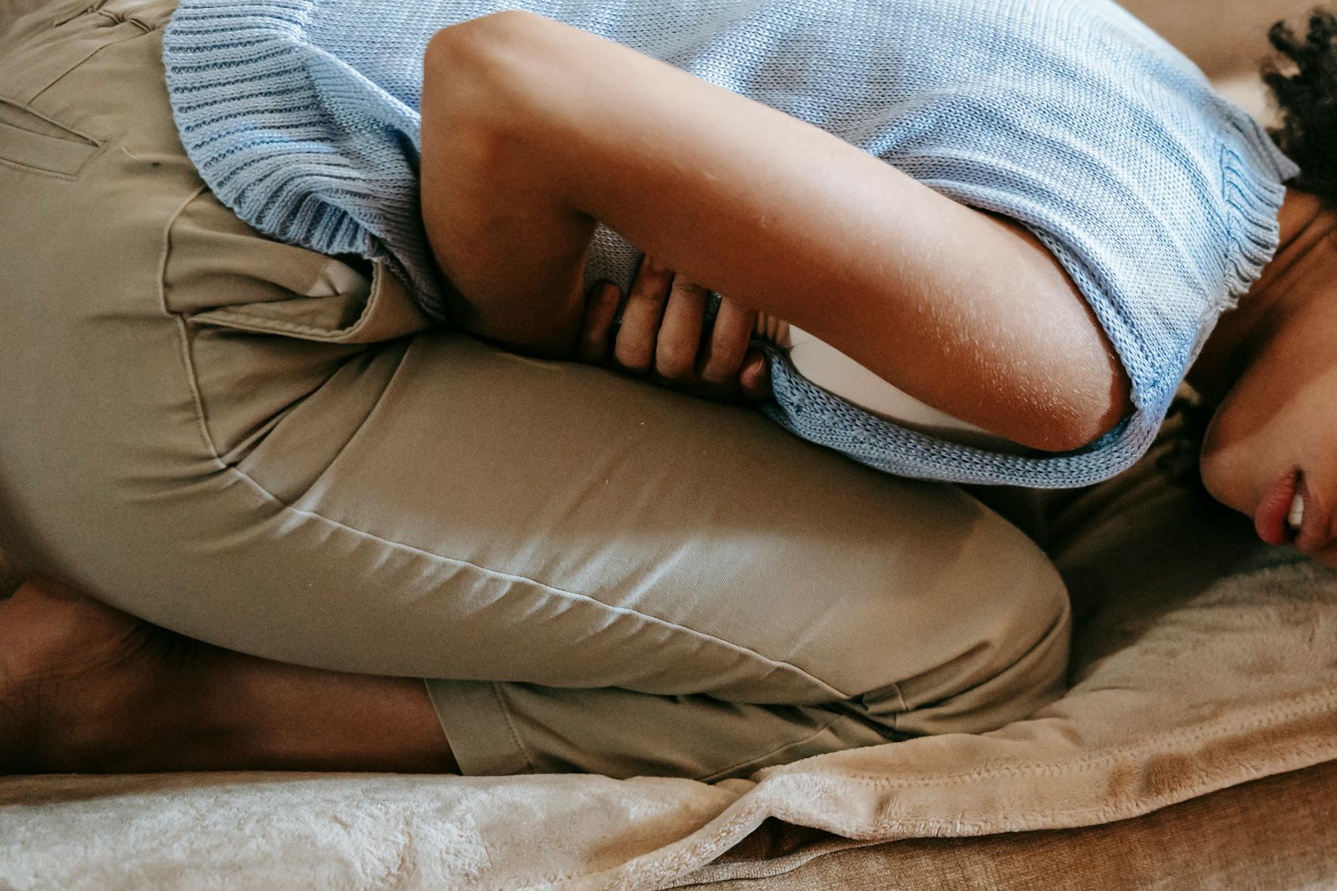 A young woman in discomfort, holding her stomach while curled up on a couch indoors.