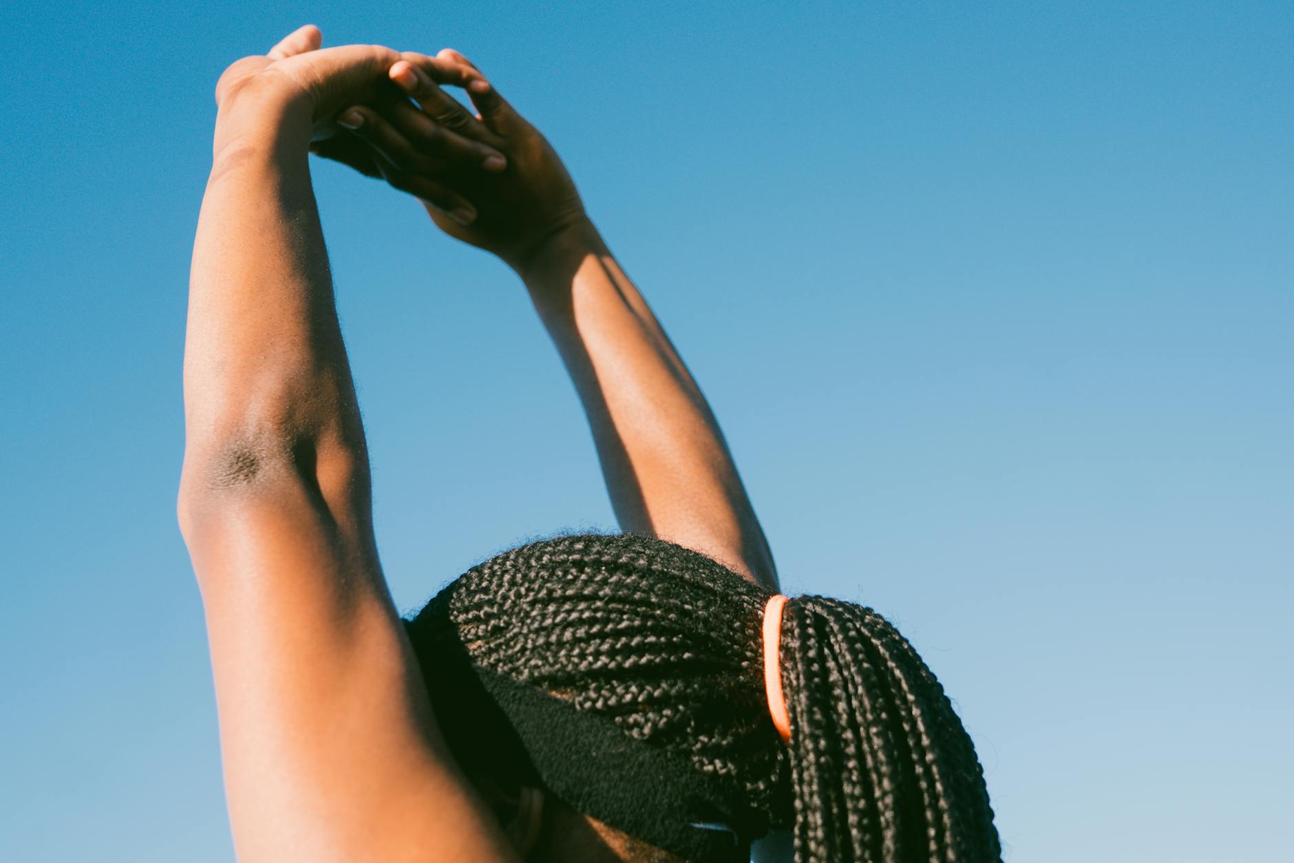 Person stretching outdoors with braided hair against a clear blue sky for a healthy lifestyle concept.